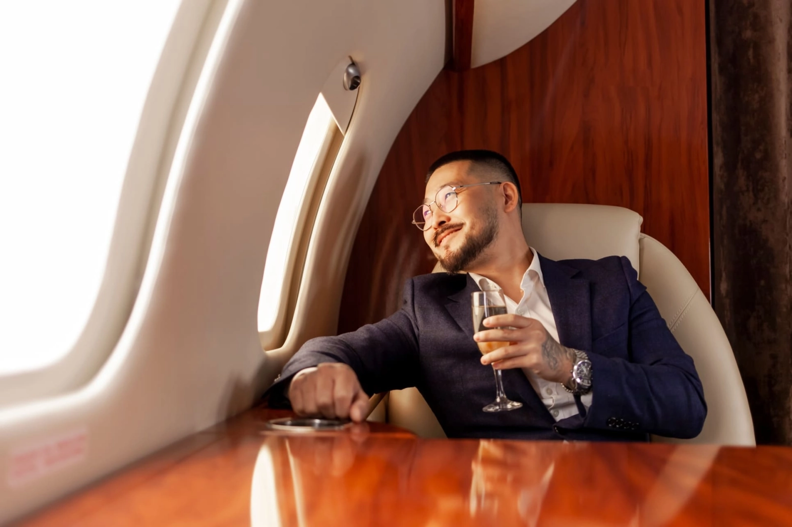 Asiatischer Geschäftsmann im Anzug und mit Brille, der mit einem Glas Champagner in einem privaten Luxusjet fliegt, koreanischer Unternehmer